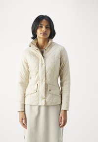 Casaco acolchoado bege com colarinho alto, botões de pressão e dois bolsos frontais. O tecido tem uma textura suave com um padrão de diamante.