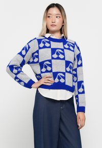 Maglione corto in morbido grigio con un audace motivo a quadri blu e design di ciliegie. I polsini e il colletto a coste aggiungono texture. Indossato sopra una camicia bianca.