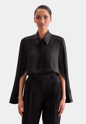 Kobieta w czarnej bluzce z rękawami w stylu peleryny oraz czarnych spodniach z wysokim stanem, stojąca na tle jednolitego jasnoszarego tła.