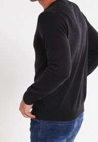 Maglione a maglia grigio scuro con maniche lunghe, texture semplice e orlo a costine. Indossato sopra jeans blu visto da un'angolazione laterale-posteriore.