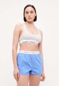 Bjelina sportska grudnjak s uzorkom i Calvin Klein logom na elasticnoj traci, uparen s svijetloplavim šortsevima s elastičnim pojasom koji prikazuju opušteni kroj.