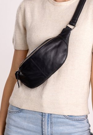 Persona che indossa un maglione lavorato a maglia beige a maniche corte e jeans azzurri chiari, con una borsa a tracolla nera in pelle sul petto.