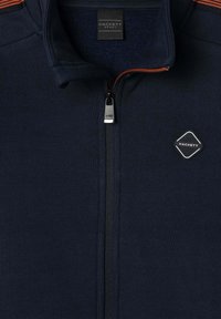 Marineblå zip-jakke med orange lynlåsforing, Hackett Sport-label indeni kraven og diamantformet Hackett-logo på brystet.