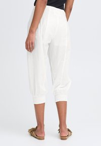 Pantalons courts blancs, légers, avec une taille smockée et des poignets élastiques. Le modèle porte des sandales à enfiler marron et présente un profil latéral.