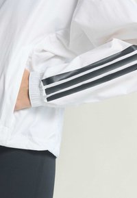 White jacket with elastic cuffs, featuring three black stripes on the sleeve. Smooth texture, lightweight material, modern design.