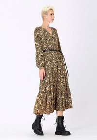 Robe noire à fleurs avec des manches longues, décolleté en V, jupe froncée et une ceinture noire, portée avec des bottes noires à lacets sur un fond clair.