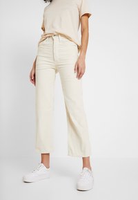 Krämsfärgade kordfloth vida byxor med hög midja, försedda med knappstängning och framfickor, tillsammans med en ljusbeige t-shirt och vita sneakers.