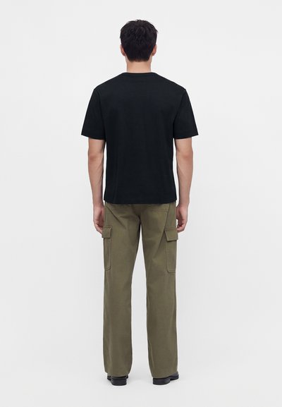 Homme portant un t-shirt noir uni à manches courtes et un pantalon cargo vert olive, debout de dos sur un fond blanc.