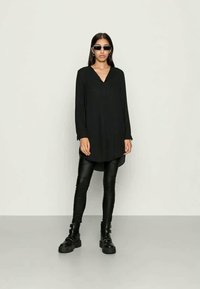 Sieviete, kura valkā melnu garu apvalku, melnas ādas bikses, masīvas melnas zābakus un taisnstūra formas melnus saulesbrilles, stāv uz vienkārša, gaiša fona.