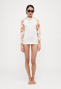 Bijela longsleeve rash guard s narančastim cvjetnim uzorcima na rukavima i "Ripcurl" logotipom na prsima. Glatka tekstura, uski dizajn.