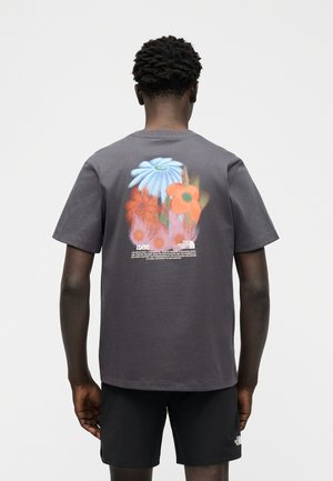 Mees, kes kannab tumehallist särki, millel on seljal värviline suur lillekujundus ja tekst, mida ta kannab mustade lühikeste pükstega.