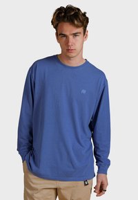 Modré tričko s dlouhým rukávem vyrobené z měkké bavlny, s kulatým výstřihem a malým vyšitým logem slona na hrudi. Má volný střih.