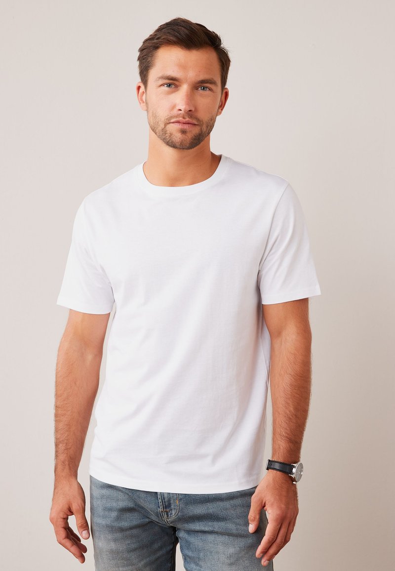 Jeune homme aux cheveux bruns courts et à la barbe porte un t-shirt blanc uni à manches courtes et un jean sur un fond neutre.