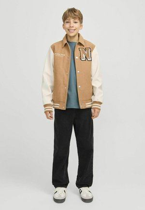 Chaqueta varsity marrón y crema con botones a presión, que presenta una gran "N" en el pecho izquierdo, usada sobre una camiseta azul petróleo y pantalones negros.
