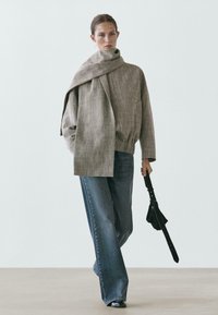 Donna con giacca grigia oversize abbinata a una sciarpa coordinata, jeans a gamba larga e scarpe nere, che tiene in mano un accessorio nero simile a una cintura.