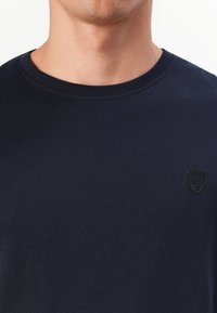 Mornarsko plava majica s kratkim rukavima, izrađena od mekog pamuka. Ima mali, izvezeni logo na lijevoj strani prsnog koša. Glatka tekstura, jednostavan dizajn.