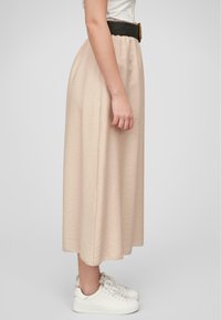 Jupe midi beige en tissu léger avec une finition texturée, associée à une large ceinture tressée noire et des baskets blanches.
