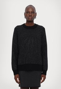 Pull en tricot noir avec un motif de pois blancs, poignets et ourlet côtelés, coupe décontractée, encolure ronde, porté sur une jupe assortie.