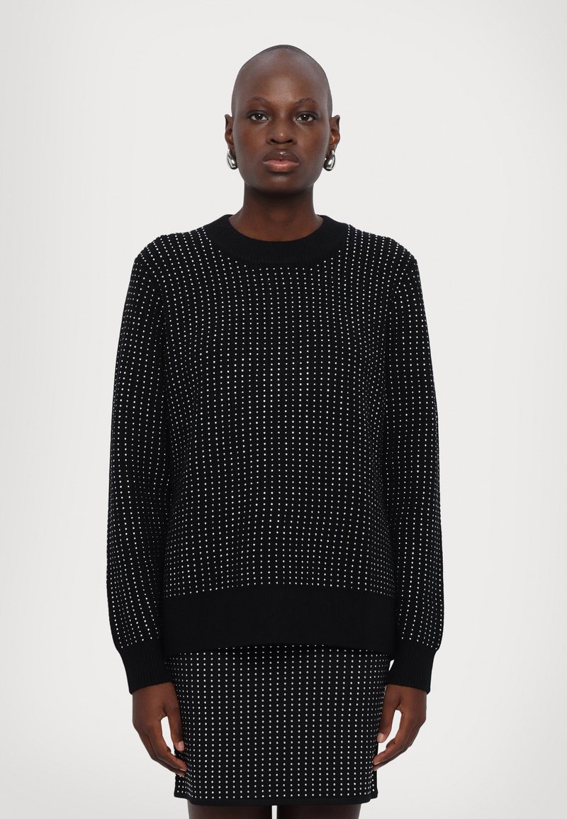 Pull en tricot noir avec un motif de pois blancs, poignets et ourlet côtelés, coupe décontractée, encolure ronde, porté sur une jupe assortie.