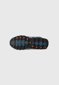 Semelle de chaussure de sport noire avec une texture en caoutchouc, présentant un design bleu et rouge. Comprend des grip circulaires et des détails de marque visibles.