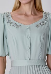 Robe vert menthe avec des manches courtes bouffantes, un décolleté en V, des cristaux décoratifs aux épaules et une jupe plissée avec une ceinture à la taille.