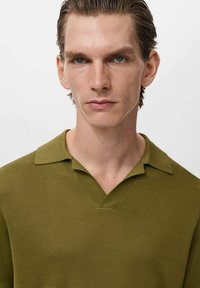 Maslinasto zelena rebrasta polo majica s V-izrezom i izrezanim ovratnikom. Kratki rukavi i teksturirani materijal daju strukturiran izgled.