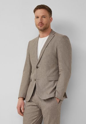 Man die een beige gestructureerd pak met wit overhemd draagt, staand met één hand in zijn zak tegen een effen lichtgrijze achtergrond.