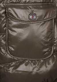 Olivgrön puffjacka i tyg, med en stor framficka som stängs med knapp och texturerade söm detaljer. Slät, glansig yta.