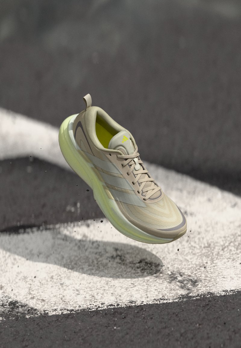 Μπεζ και κίτρινο παπούτσι για τρέξιμο Adidas στον αέρα πάνω από άσφαλτο με μια λευκή βαμμένη γραμμή, ρίχνοντας σκιά και ανυψώνοντας μικρά χαλίκια.