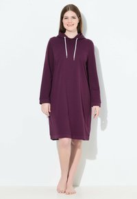 Junge Frau steht barfuß und trägt ein lila Kapuzenkleid mit langen Ärmeln und knie-langer Länge, versehen mit weißen Kordeln, und lächelt vor einem weißen Hintergrund.