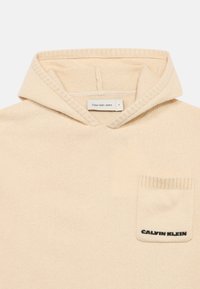 Μπέζ φούτερ από μαλακό ύφασμα με τσέπη καγκουρό, ριμπ ύφανση και ετικέτα που αναγράφει "Calvin Klein Jeans" στην τσέπη του στήθους.