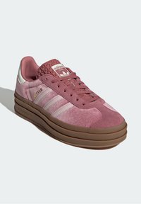 Růžová teniska Adidas Gazelle s světle růžovými pruhy, texturovaným jazykem, hnědou podrážkou a logem Adidas na boku a jazyku.