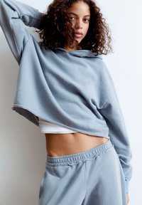 Haut de survêtement gris clair à capuche avec cordon, texture douce et manches longues. Assorti à un pantalon de survêtement avec une taille élastique.