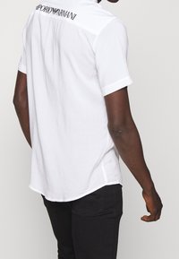 Man med kortärmad vit Emporio Armani-skjorta med logotyp på övre ryggen, ihop med svarta byxor, vänd bort från kameran.