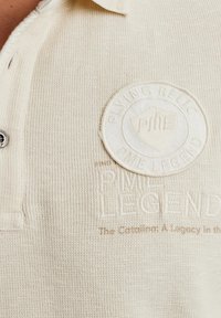 Polo texturé couleur crème avec un col, présentant un écusson rond en relief et un logo, ainsi que du texte brodé sous l'écusson.