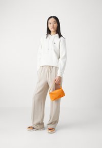 Felpa bianca con cappuccio e lacci, pantaloni beige a gamba larga e texturizzati, borsa arancione e sneakers multicolori con suole spesse.