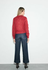 Červený sveter s pleteným vzorom a textúrovaným chrbtom, ktorý je skombinovaný s tmavomodrými širokými džínmi. Oblečenie dopĺňajú členkové topánky na podpätku.