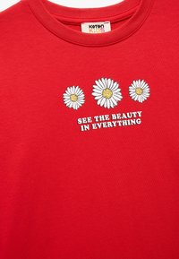 Röd bomulls-t-shirt med tre vita tusenskönor och texten "SE SKÖNHETEN I ALLT" tryckt i vitt och svart.