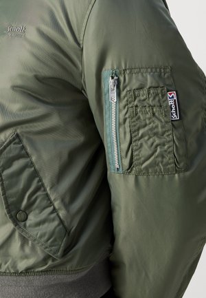 Chaqueta bomber verde con una textura suave y brillante, que cuenta con un bolsillo con cremallera en la manga izquierda y un parche de logo visible.