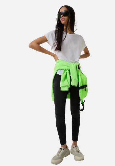 Hvid t-shirt, neongrøn hættetrøje bundet om livet, sorte leggings og beige sneakers. Sorte oversized solbriller fuldender looket.