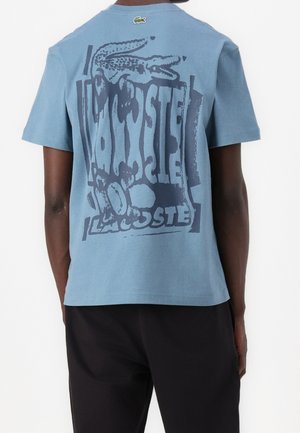 Mand iført lyseblå kortærmet T-shirt med stort mørkeblått forvrænget Lacoste-logo og krokodillegrafik på ryggen.