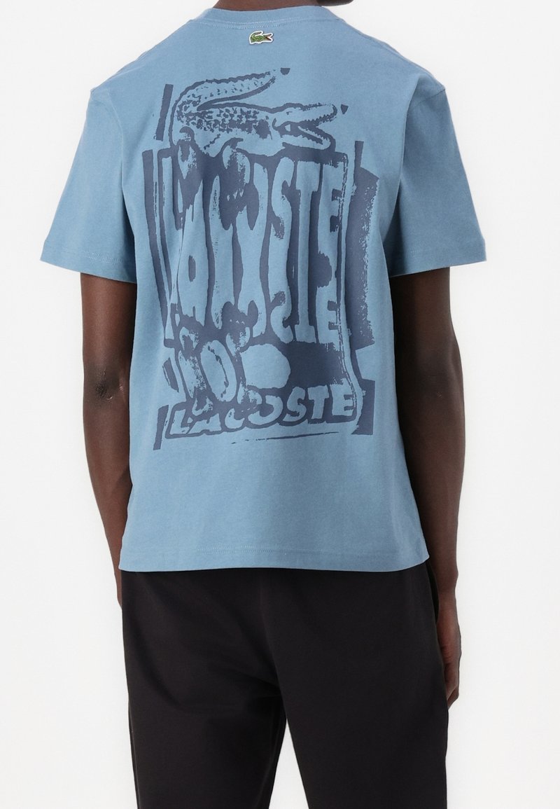Homme portant un T-shirt à manches courtes bleu clair avec un grand logo Lacoste déformé bleu foncé et un graphisme de crocodile dans le dos.