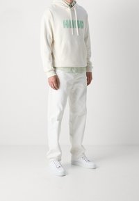 Felpa crema con testo "HUGO" verde, indossata sopra una maglietta verde chiaro; abbinata a jeans bianchi e sneakers bianche. Tessuto morbido e vestibilità casual.