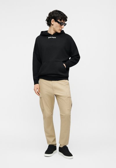 Mees, kes kannab musta "Palm Angels" kapuutsi, beežide cargo-pükse, musti tossusid ja musti päikeseprille, seisab ühe käega taskus.