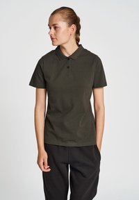 Donkergroen polo met een kraag, twee knopen, en korte mouwen, gedragen met losse zwarte broek. Een gladde katoenstof.