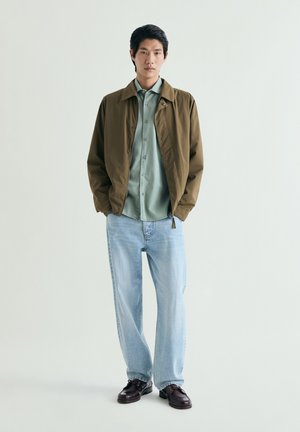 Joven de pie, vistiendo chaqueta marrón, camisa de botones verde claro, vaqueros azul claro y zapatos marrón oscuro, sobre un fondo liso.