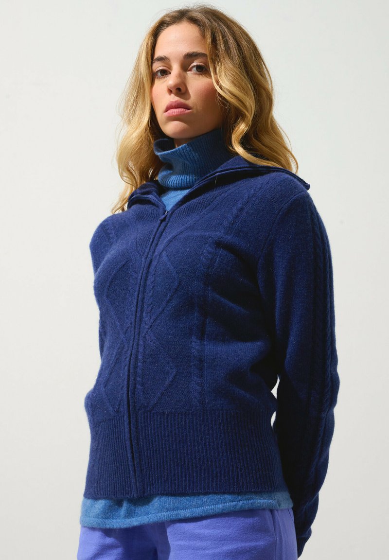 Mörkblå zip-up ulltröja med ett strukturerat diamantmönster, med en hög krage. Under finns en ljusblå polotröja synlig.