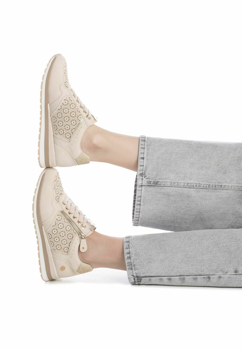 Zapatillas de piel perforada en color beige con diseño de cordones y detalle de cremallera, combinadas con jeans de pierna ancha en color gris.