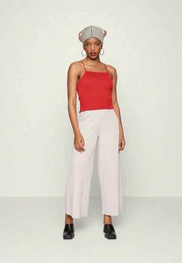 Mulher vestindo uma blusa vermelha sem mangas, calças largas beije claro, sapatos pretos, lenço listrado na cabeça e brincos grandes em forma de argola, em pé sobre um fundo simples.