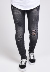 Zwarte distressed jeans met meerdere scheuren, spetters en een slim fit. Gecombineerd met zwart-witte low-top sneakers.